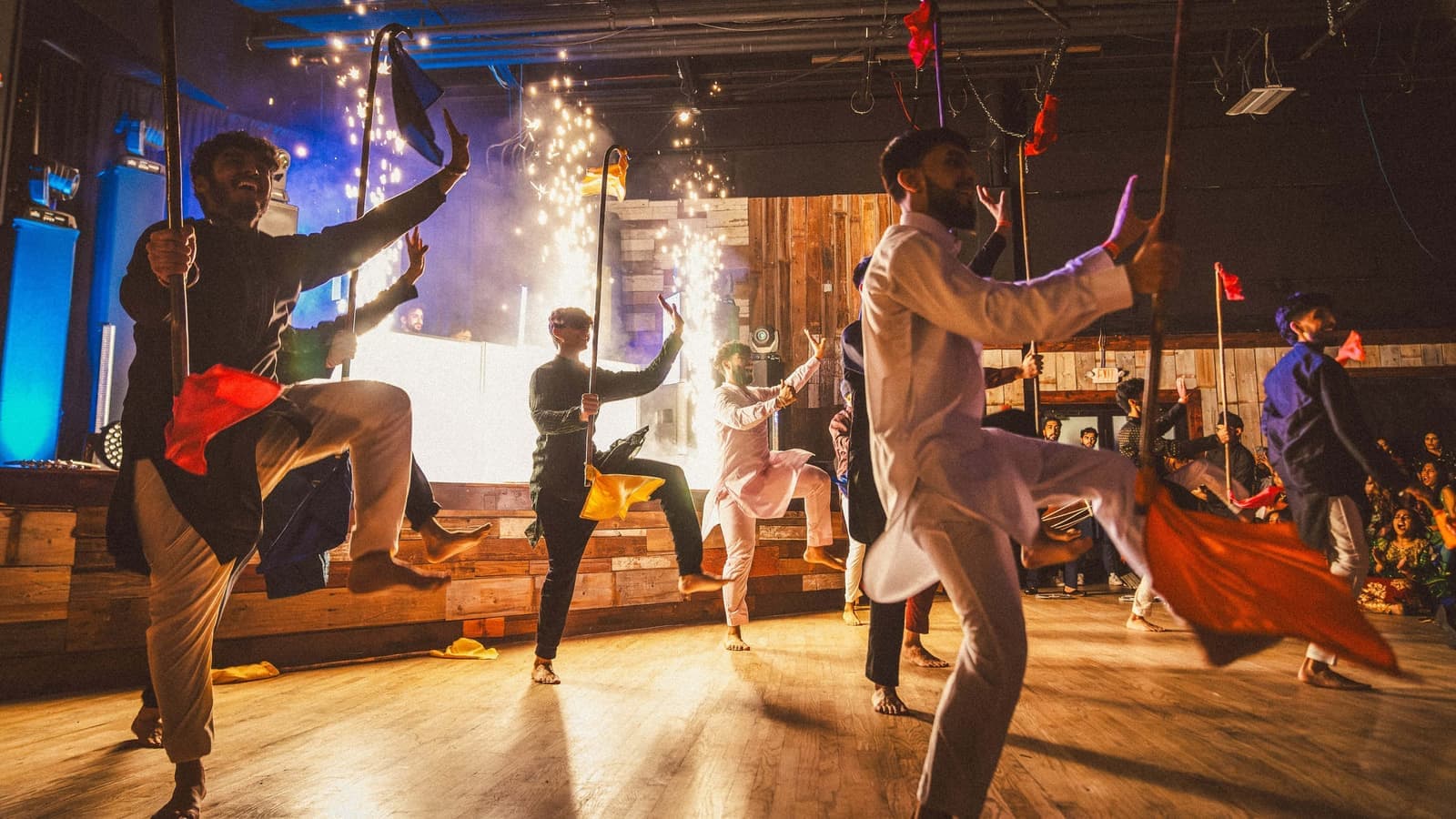 Performers mid-air during a bhangra flag dance with stage sparks behind them